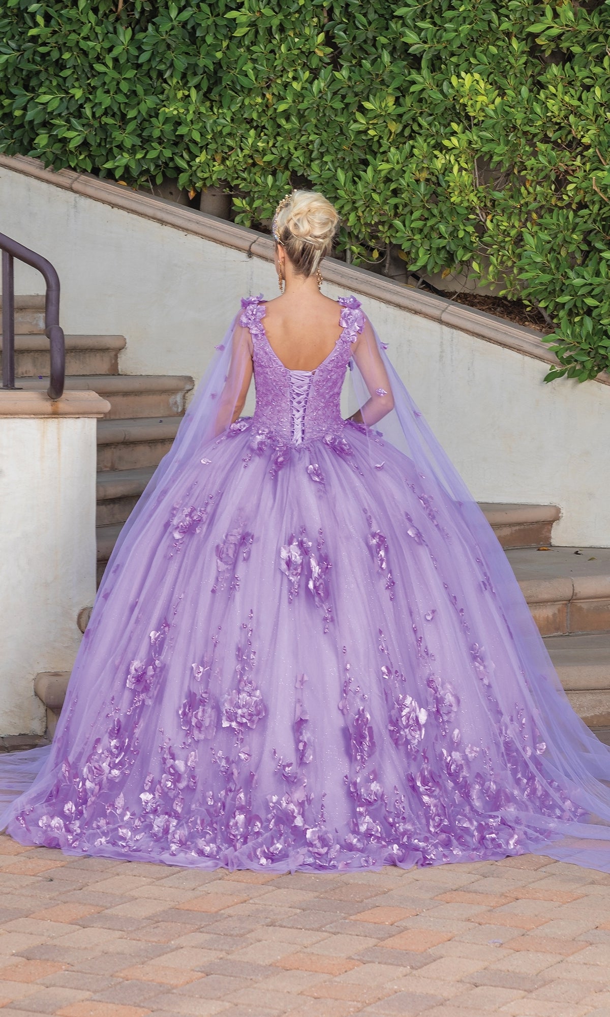 Wearing the Quinceanera Dress 1695 By Dancing Queen, a person in an a-line gown with floral details and sheer cape sleeves stands on a patio facing away, steps and greenery behind them.